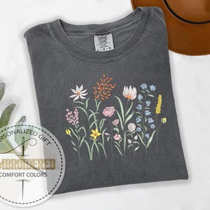 May include: A dark gray t-shirt with an embroidered design of colorful flowers. The flowers are in shades of pink, yellow, blue, orange, and white. The t-shirt has a crew neck and short sleeves.