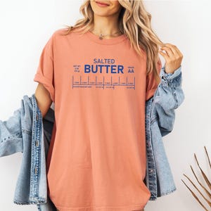 May include: A coral-coloured t-shirt with the words "SALTED BUTTER" in blue block letters. Below the text are blue lines indicating measurements. The shirt is worn with a denim jacket.