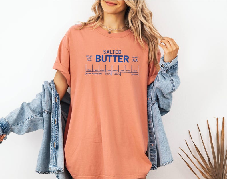 May include: A coral-coloured t-shirt with the words "SALTED BUTTER" in blue block letters. Below the text are blue lines indicating measurements. The shirt is worn with a denim jacket.