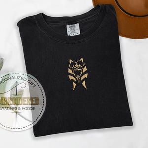 May include: Black t-shirt with a gold embroidered design of a stylised wolf head with a skull in the centre. The design is on the front of the shirt, centred on the chest. The shirt is folded and laid flat on a white surface.