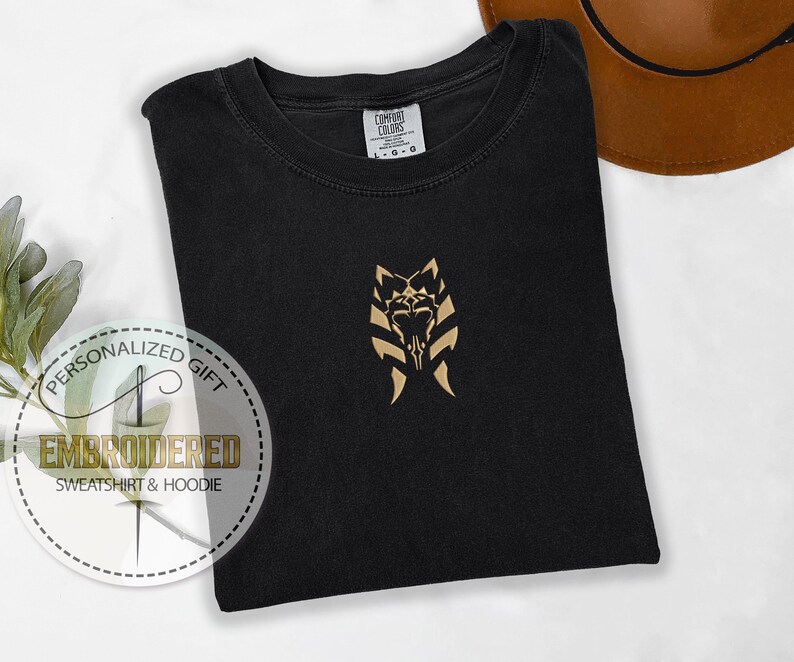 May include: Black t-shirt with a gold embroidered design of a stylised wolf head with a skull in the centre. The design is on the front of the shirt, centred on the chest. The shirt is folded and laid flat on a white surface.