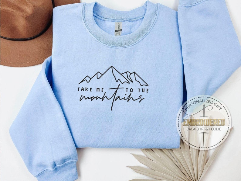 K&ouml;nnte beinhalten: Hellblaues Sweatshirt mit dem schwarzen Text "TAKE ME TO THE mountains". Das Design zeigt eine Bergkette. Im Hintergrund ist ein brauner Hut zu sehen.