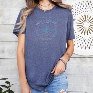 K&ouml;nnte beinhalten: Ein blaues T-Shirt mit einem grafischen Design, das "Ode to Storm" und "Brave the Dark" mit einem stilisierten Auge und einem floralen Design sagt.