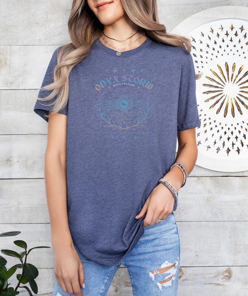 K&ouml;nnte beinhalten: Ein blaues T-Shirt mit einem grafischen Design, das "Ode to Storm" und "Brave the Dark" mit einem stilisierten Auge und einem floralen Design sagt.