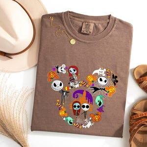 May include: A brown t-shirt with a graphic print of characters from the Nightmare Before Christmas movie. The characters are arranged in a heart shape with a Mickey Mouse head outline. The graphic includes Jack Skellington, Sally, and other characters from the movie.