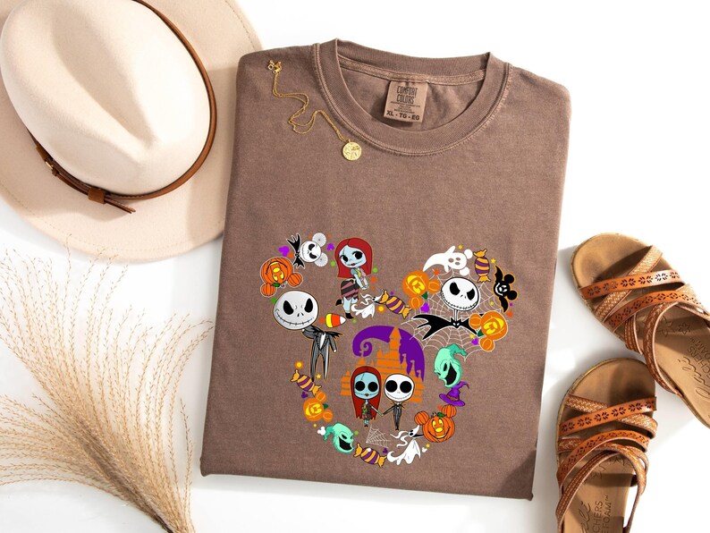 May include: A brown t-shirt with a graphic print of characters from the Nightmare Before Christmas movie. The characters are arranged in a heart shape with a Mickey Mouse head outline. The graphic includes Jack Skellington, Sally, and other characters from the movie.