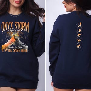 May include: Navy blue sweatshirt with a graphic print of a mountain range and the text "ONYX STORM - BRAVE THE DARK - I WILL NOT DIE TODAY I WILL SAVE HIM". The back of the sweatshirt features a vertical row of five orange dragon silhouettes.