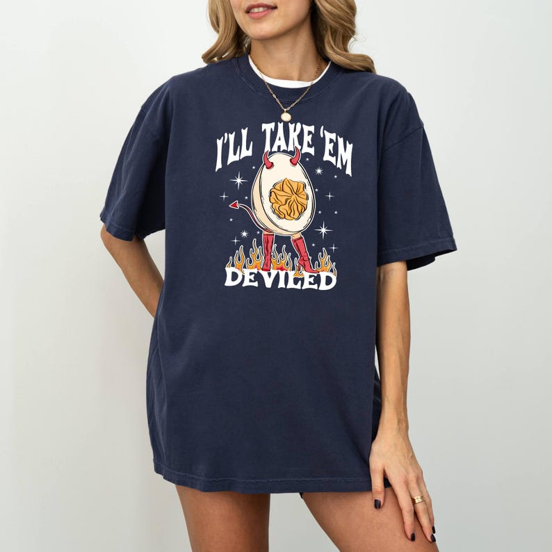 以下が含まれることがあります： 悪魔の卵のグラフィックが描かれたネイビーブルーのTシャツ。卵は角と赤い靴を履いています。Tシャツには「I'll take 'em deviled」という文字が印刷されています。