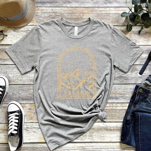 May include: A heather gray t-shirt with a gold graphic design featuring mountains and the text "Vellaris". The shirt is paired with blue jeans, black sneakers, a straw hat, and a watch. The items are arranged on a wooden surface.