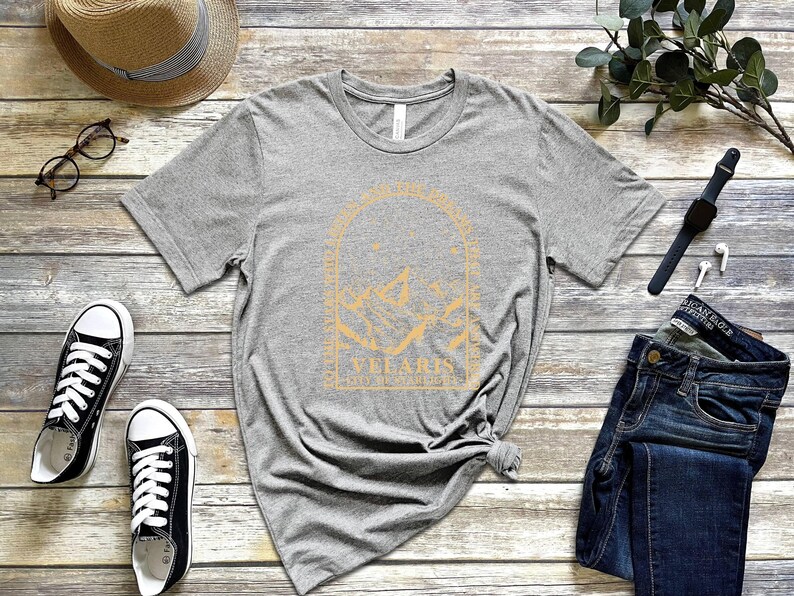 May include: A heather gray t-shirt with a gold graphic design featuring mountains and the text "Vellaris". The shirt is paired with blue jeans, black sneakers, a straw hat, and a watch. The items are arranged on a wooden surface.