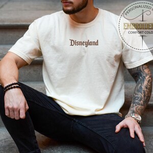 Pu&ograve; includere: T-shirt color crema con la scritta "Disneyland" ricamata in marrone. La maglietta ha maniche corte e girocollo. L'immagine include anche una grafica circolare con le parole "Regalo Personalizzato" e "Comfort Colors Ricamato".