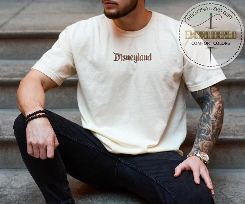 Pu&ograve; includere: T-shirt color crema con la scritta "Disneyland" ricamata in marrone. La maglietta ha maniche corte e girocollo. L'immagine include anche una grafica circolare con le parole "Regalo Personalizzato" e "Comfort Colors Ricamato".