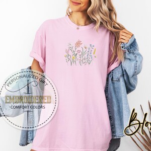 May include: A pink t-shirt with an embroidered floral design. The design features a variety of flowers in shades of pink, yellow, blue, and green.