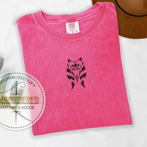 May include: A pink t-shirt with a black embroidered design of a Mandalorian symbol. The t-shirt is folded and laid flat on a white surface. The text "PERSONALIZED GIFT EMBROIDERED SWEATSHIRT & HOODIE" is visible in a circle on the left side of the image.