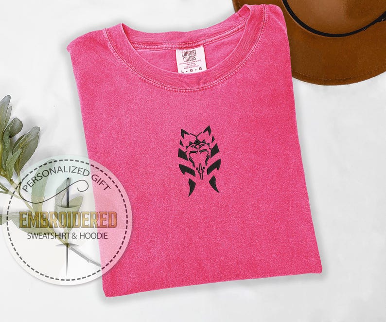 May include: A pink t-shirt with a black embroidered design of a Mandalorian symbol. The t-shirt is folded and laid flat on a white surface. The text "PERSONALIZED GIFT EMBROIDERED SWEATSHIRT & HOODIE" is visible in a circle on the left side of the image.
