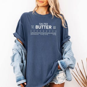May include: A navy blue t-shirt featuring the words "SALTED BUTTER" in white, with a graphic showing butter measurements. The shirt also includes the text "NET WT. 113 g" and "GRADE AA".