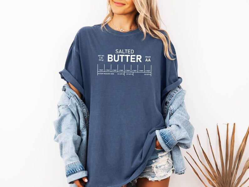 May include: A navy blue t-shirt featuring the words "SALTED BUTTER" in white, with a graphic showing butter measurements. The shirt also includes the text "NET WT. 113 g" and "GRADE AA".