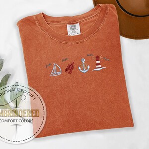 May include: Burnt orange Comfort Colors t-shirt with embroidered nautical designs: sailboat, lobster, anchor, and lighthouse. The shirt has a crew neck and short sleeves. The shirt is folded and has a personalized gift label.