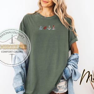 May include: Moss green t-shirt with embroidered nautical designs: a sailboat, a red buoy, an anchor, and a lighthouse. The shirt has rolled-up sleeves and the text "Personalized Gift" and "Embroidered Comfort Colors" in a circular design.