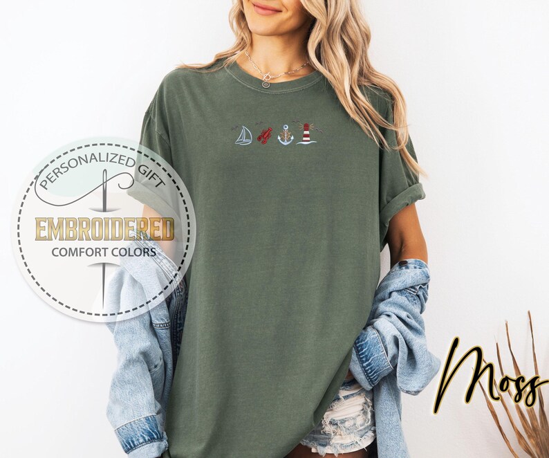 May include: Moss green t-shirt with embroidered nautical designs: a sailboat, a red buoy, an anchor, and a lighthouse. The shirt has rolled-up sleeves and the text "Personalized Gift" and "Embroidered Comfort Colors" in a circular design.