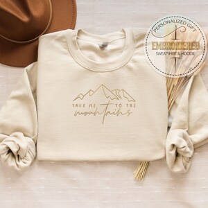 K&ouml;nnte beinhalten: Cremefarbenes Sweatshirt mit dem Schriftzug "Take me to the mountains" und einer goldenen Berg-Grafik. Das lang&auml;rmlige Sweatshirt hat einen Rundhalsausschnitt. Ein brauner Hut ist im Hintergrund zu sehen.