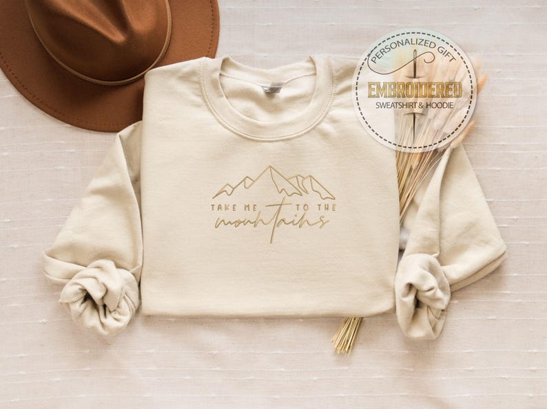 K&ouml;nnte beinhalten: Cremefarbenes Sweatshirt mit dem Schriftzug "Take me to the mountains" und einer goldenen Berg-Grafik. Das lang&auml;rmlige Sweatshirt hat einen Rundhalsausschnitt. Ein brauner Hut ist im Hintergrund zu sehen.