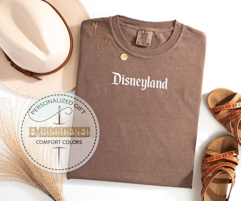 Pu&ograve; includere: Una maglietta marrone con la scritta "Disneyland" in bianco. Una collana d'oro e sandali marroni sono accanto alla maglietta. Un cappello beige con una fascia marrone e una toppa ricamata sono anche nell'immagine.