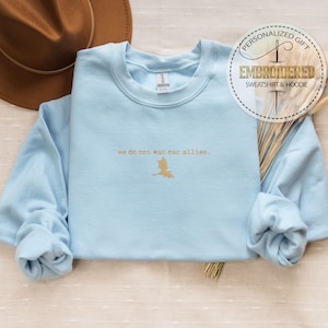 May include: Light blue sweatshirt with a gold embroidered design that says "we do not eat our allies."