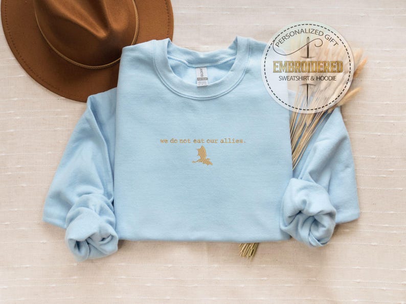 May include: Light blue sweatshirt with a gold embroidered design that says "we do not eat our allies."