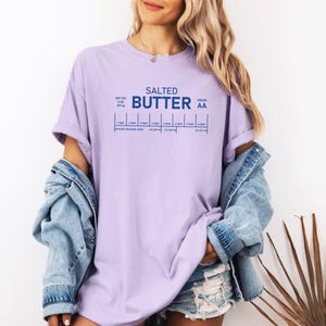 May include: A lilac t-shirt featuring the words "Salted Butter" in blue, with a graphic showing measurement conversions. The shirt is made of a soft, comfortable material.