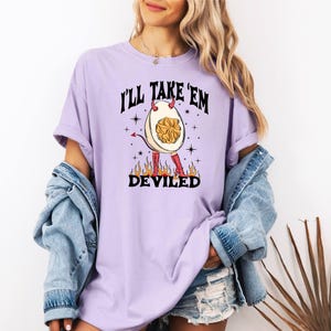 以下が含まれることがあります： 悪魔の卵のグラフィックが描かれた紫色のTシャツ。角と尻尾があります。シャツのテキストは「I'll Take 'Em Deviled」です。