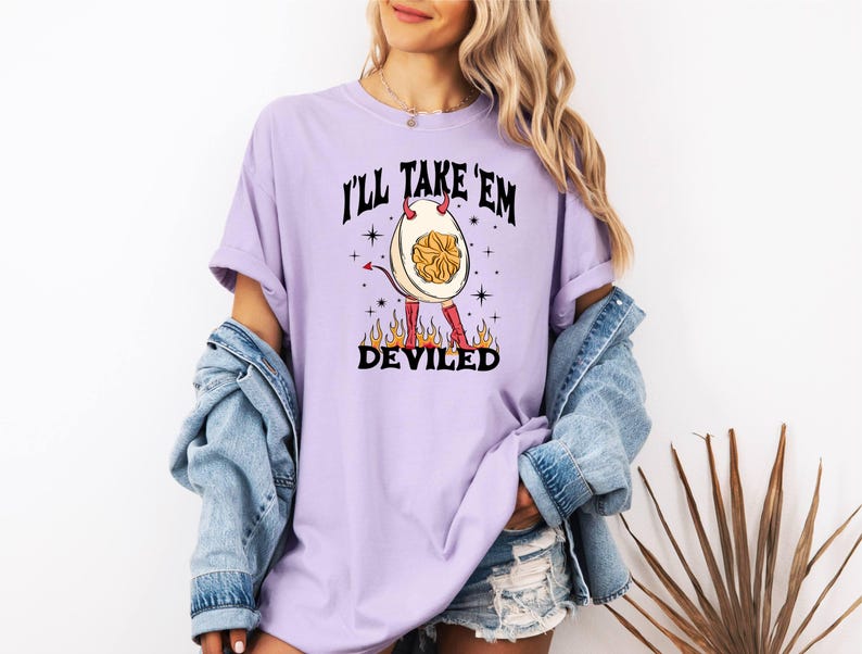 以下が含まれることがあります： 悪魔の卵のグラフィックが描かれた紫色のTシャツ。角と尻尾があります。シャツのテキストは「I'll Take 'Em Deviled」です。