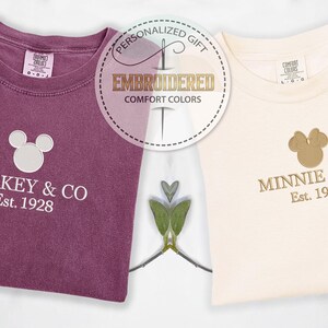 May include: Two embroidered Comfort Colors t-shirts. One is a deep purple with "Mickey & Co Est. 1928" and a Mickey Mouse head. The other is cream-colored with "Minnie & Co Est. 1928" and a Minnie Mouse head.
