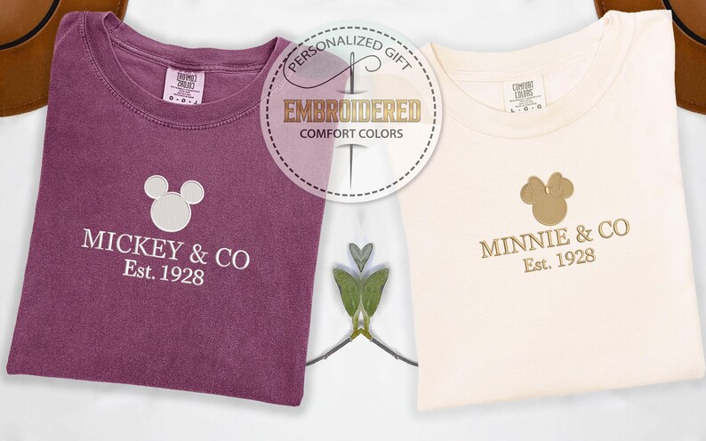May include: Two embroidered Comfort Colors t-shirts. One is a deep purple with "Mickey & Co Est. 1928" and a Mickey Mouse head. The other is cream-colored with "Minnie & Co Est. 1928" and a Minnie Mouse head.