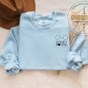 May include: A light blue sweatshirt with the text "DISNEY BOUND" and a Mickey Mouse head design. The sweatshirt is embroidered and has long sleeves. A brown hat is in the background. The sweatshirt is a personalized gift.