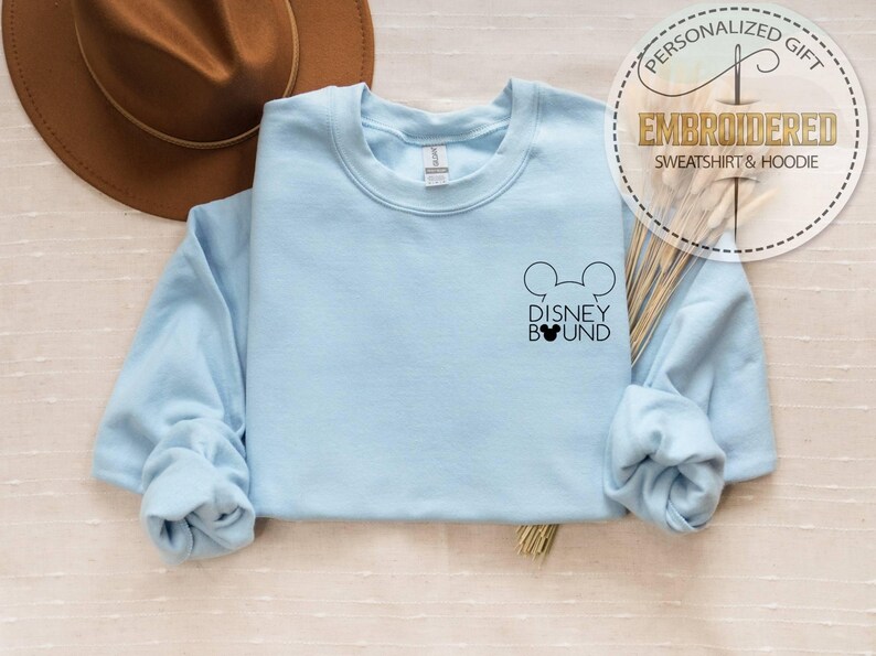 May include: A light blue sweatshirt with the text "DISNEY BOUND" and a Mickey Mouse head design. The sweatshirt is embroidered and has long sleeves. A brown hat is in the background. The sweatshirt is a personalized gift.