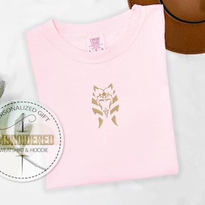 May include: A pink t-shirt featuring a gold embroidered design of a Mandalorian symbol. The t-shirt has a tag that reads "Comfort Colors".