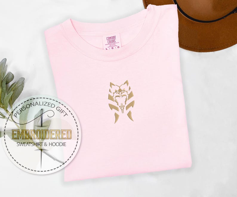 May include: A pink t-shirt featuring a gold embroidered design of a Mandalorian symbol. The t-shirt has a tag that reads "Comfort Colors".