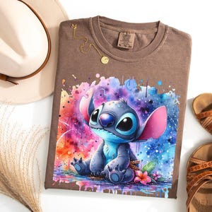 May include: Brown t-shirt with a watercolour illustration of Stitch from Lilo & Stitch. The design features vibrant colours and a splash effect. A gold necklace and a beige hat are also visible.
