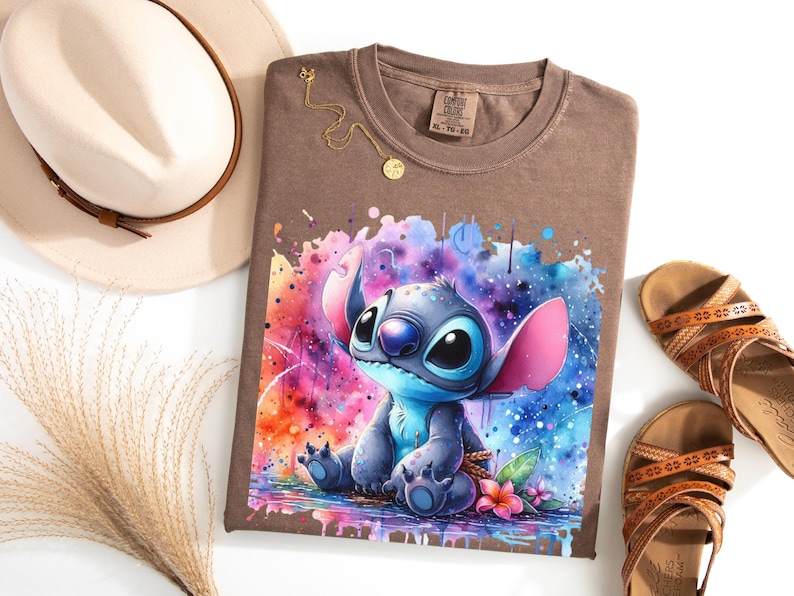 May include: Brown t-shirt with a watercolour illustration of Stitch from Lilo & Stitch. The design features vibrant colours and a splash effect. A gold necklace and a beige hat are also visible.