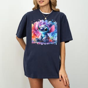 May include: Navy blue t-shirt with a colourful watercolour graphic of Stitch from Lilo & Stitch. The design shows Stitch sitting with a vibrant splash of colours behind him. The shirt has a comfortable, oversized fit.