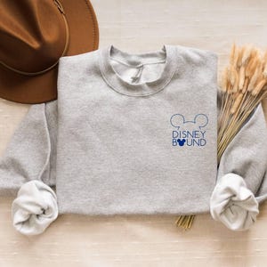 May include: A heather gray sweatshirt with the words "DISNEY BOUND" and Mickey Mouse ears in navy blue. A brown hat and dried wheat stalks are also in the image. The sweatshirt has rolled-up sleeves.