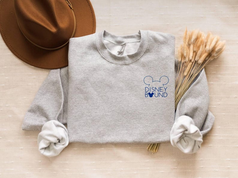 May include: A heather gray sweatshirt with the words "DISNEY BOUND" and Mickey Mouse ears in navy blue. A brown hat and dried wheat stalks are also in the image. The sweatshirt has rolled-up sleeves.