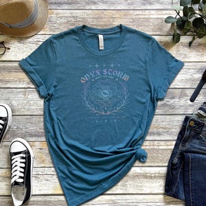 K&ouml;nnte beinhalten: Ein blaues T-Shirt mit einem grafischen Design eines Blumenkranzes mit dem Text "Onyx Storm Save the Fire" in rosa und lila Schrift.