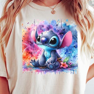 May include: A cream-coloured t-shirt with a watercolour illustration of the character Stitch from Lilo & Stitch. The artwork depicts Stitch in blue tones, with pink ears, set against a vibrant, colourful background with paint splashes and flowers.