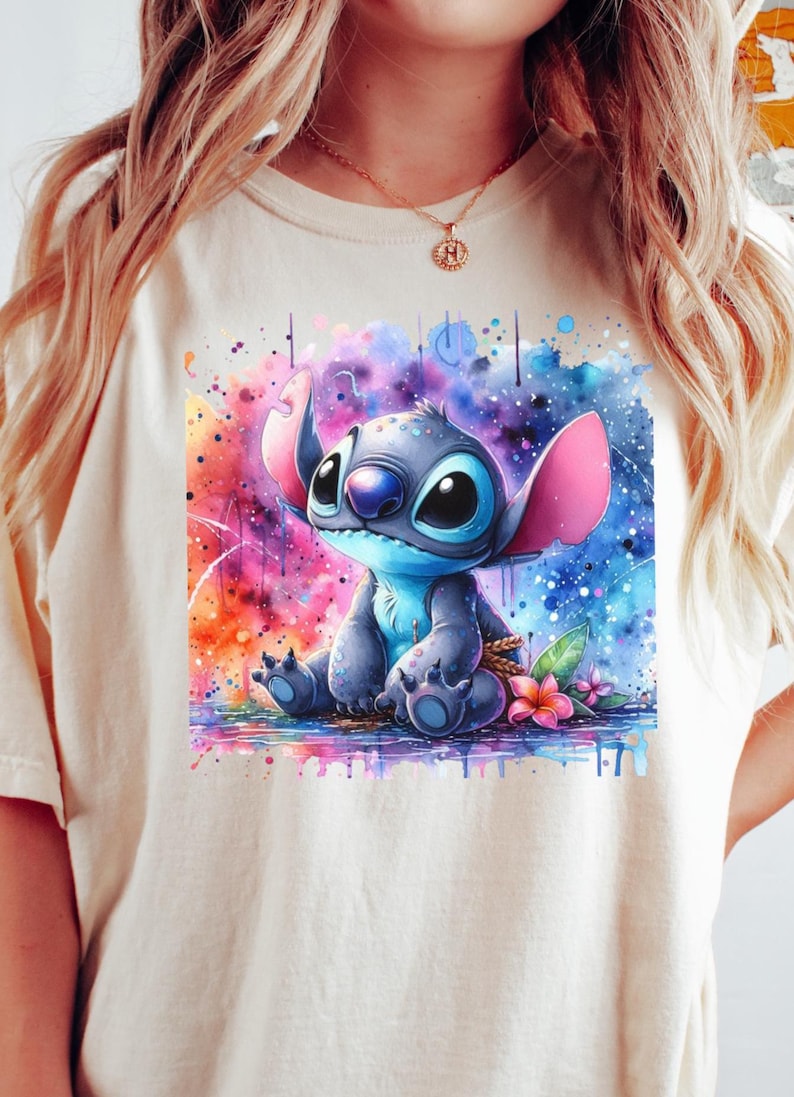 May include: A cream-coloured t-shirt with a watercolour illustration of the character Stitch from Lilo & Stitch. The artwork depicts Stitch in blue tones, with pink ears, set against a vibrant, colourful background with paint splashes and flowers.