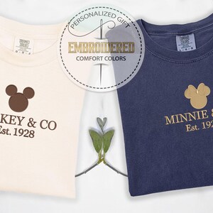 May include: Two folded t-shirts, one cream and one navy blue, with embroidered logos. The cream shirt says "Mickey & Co. Est. 1928" in brown. The navy shirt says "Minnie & Co. Est. 1928" in gold. A circular graphic reads "Embroidered Comfort Colors".