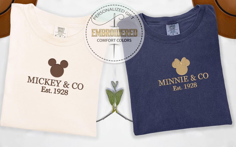 May include: Two folded t-shirts, one cream and one navy blue, with embroidered logos. The cream shirt says "Mickey & Co. Est. 1928" in brown. The navy shirt says "Minnie & Co. Est. 1928" in gold. A circular graphic reads "Embroidered Comfort Colors".