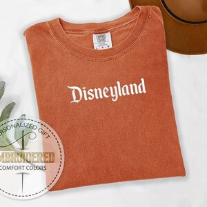 Pu&ograve; includere: Una maglietta arancione Comfort Colors con la scritta "Disneyland" ricamata in corsivo bianco. La maglietta &egrave; piegata e appoggiata su una superficie bianca. Un cappello marrone &egrave; visibile nell'angolo in alto a destra. Un adesivo regalo personalizzato &egrave; nell'angolo in basso a sinistra.