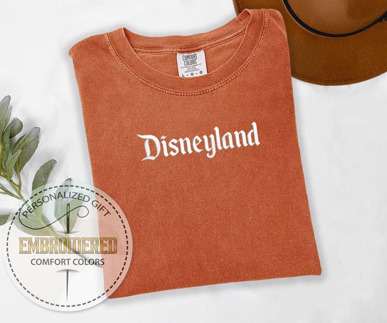 Pu&ograve; includere: Una maglietta arancione Comfort Colors con la scritta "Disneyland" ricamata in corsivo bianco. La maglietta &egrave; piegata e appoggiata su una superficie bianca. Un cappello marrone &egrave; visibile nell'angolo in alto a destra. Un adesivo regalo personalizzato &egrave; nell'angolo in basso a sinistra.